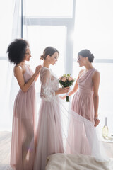 Naklejka premium pretty bride holding wedding bouquet while interracial bridesmaids adjusting her dress on sunny morning.