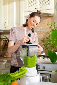 happy and cheerful caucasian woman in a t-shirt in her kitchen makes a green smoothie from fresh fruits and vegetables, a healthy vegan and vegetarian drink for health and proper nutrition after