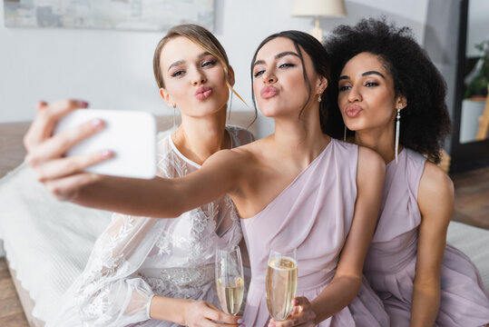 pretty bride with multicultural bridesmaids blowing air kisses while taking selfie on blurred foreground.