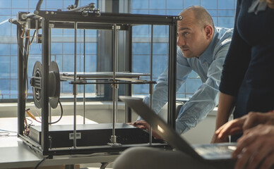 
An engineer set the 3D printer and components in the lab while his colleagues watch a sketch of the product on a laptop.Technology and engineering concept