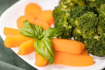 healthy food, boiled broccoli, carrots and basal leaf on a white plate on a dark green cloth