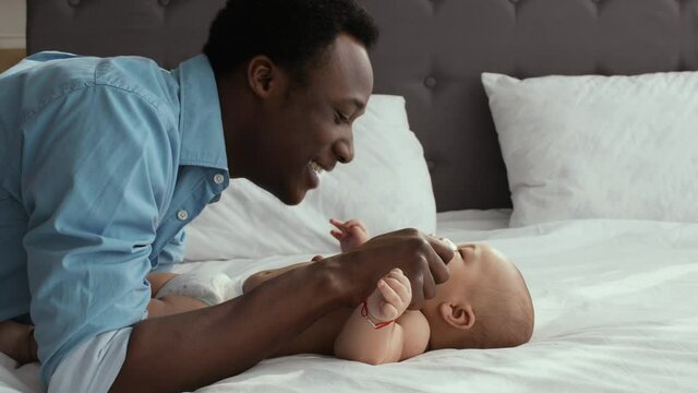 Paternity leave. Happy african american man playing with his cute newborn baby lying on big bed, smiling and laughing