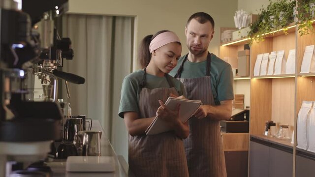 Lockdown of middle-aged Caucasian man and young mixed-race woman wearing uniform standing together in cafe and making report using notepad woman holding in hands