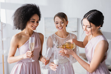 pretty woman pouring champagne near bride and african american bridesmaid.
