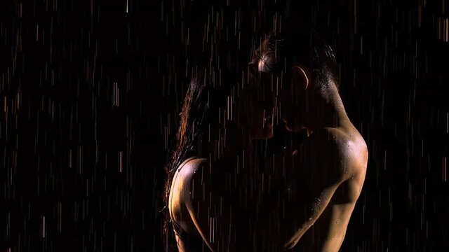 Passionate young couple dancing and enjoying pouring rain at night. Silhouettes of naked bodies in drops of water. Man gently caresses his girlfriend. Close up. Slow motion.