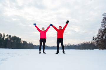 Smiling mature couple standing in the middle of frozen pond with outstretched arms in snowy winter forest. Active lifestyle concept.