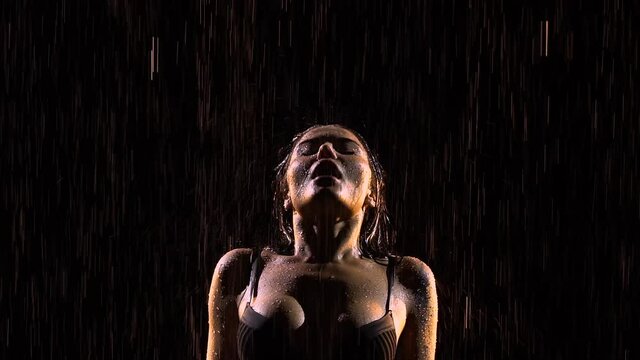 Sensual woman creates splashes of hair and strokes body with hands. Beautiful sexy brunette posing in rain. Silhouette of young sexy female body in underwear in drops of water. Close up. Slow motion.