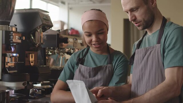 Tilt up of middle-aged Caucasian male barista standing at worktop in cafe and explaining new female mixed-race colleague how to choose necessary coffee typeTilt up of middle-aged Caucasian male barist