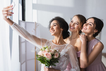 young african american bride taking selfie with cheerful bridesmaids at home. - Powered by Adobe