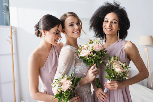 pretty bride together with interracial bridesmaids holding wedding bouquets in bedroom.