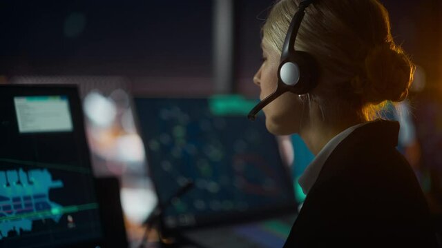Close Up Female Portrait of Air Traffic Controller with Headset Talk on a Call in Airport Tower at Night. Office Room is Full of Computer Displays with Navigation Screens, Airplane Flight Radar Data.