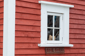 A small closed glass window with wide white trim. The exterior wall of the building is a bright red narrow wood clapboard. There's a sign with the words she shed under the window ledge.