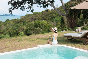 Obraz premium Happy woman in white swimsuit swimming in luxury pool hotel against beautiful view, young female with hat enjoy in tropical resort. Relaxing, summer travel, holiday and vacation concept