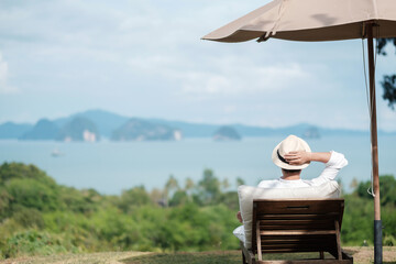 Happy traveler in white shirt and hat enjoy beautiful sea view, young man sitting on chair and looking ocean at tropical beach. Freedom, relaxing, vacation holiday and summer travel concept