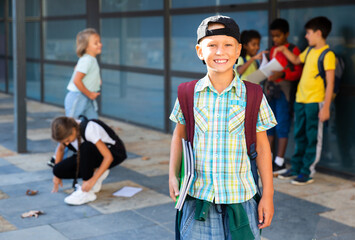 Smiling primary school boy standing outdoors, his friends talking and having fun on background