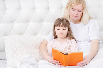 Mom reading a knuzh to a girl with down syndrome in the bedroom on the bed as she goes to bed. Ordinary childcare in a family for children with disabilities