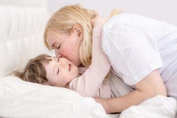 Mom kissing a girl with down syndrome in the bedroom on the bed while going to bed. Ordinary childcare in a family for children with disabilities