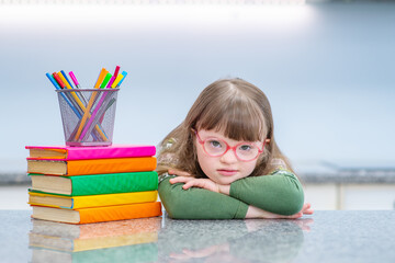 A girl with down syndrome sitting at a table at home, leaning on a stack of books that are in front of her, looking at the camera with smile