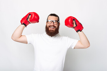Portrait of young bearded man in white t-shirt celebrating victory and wearing red boxing gloves