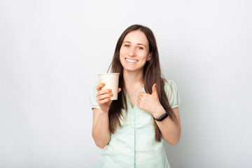Photo of happy young smiling woman holding cup of coffee to go and showing thumb up