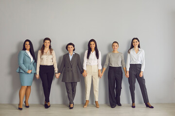 Women business team, cooperation, working in office. Team of young positive businesswomen corporate partners employees standing and holding hands together looking at camera over grey wall background