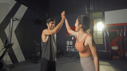 A Caucasian man and Asian woman giving High five at training workout gym fitness center club. Exercise indoor with sport equipment. People lifestyle recreation. Successful team.