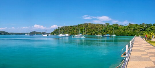 Panoramic view of beautiful Muelle Bay in the tourist resort of Puerto Galera, Philippines, with yachts moored in tranquil turquoise water, while a public promenade and park are visible on the right.