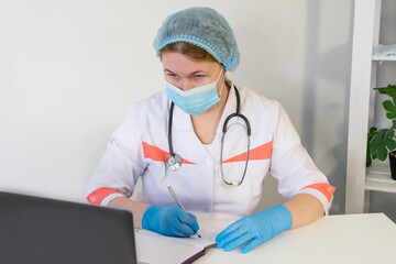 A female nurse, wearing a protective mask and blue medical gloves, works at a laptop, she enters the patient's data.