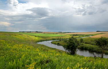 Summer cloudy landscape with dark stormy clouds over the calm river and green meadows with blooming wildflowers.