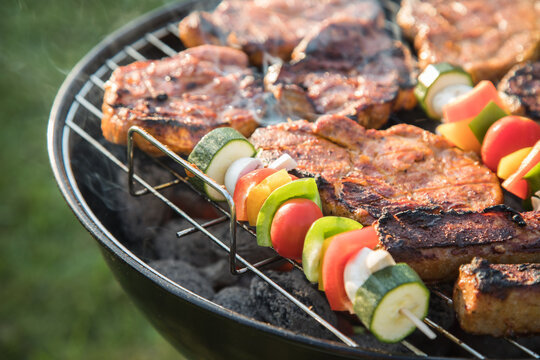 Marinierte Schweine Fleisch Nackensteaks und bunte Gemüsespieße grillen mit Rauch auf Holzkohle Kugelgrill im Garten an Sommer Abend