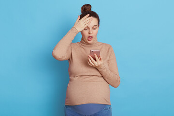 Obraz premium Shocked pregnant female wearing beige turtleneck looking a t smart phone screen with opened mouth, touching her head, sees breaking news, isolated over blue background.
