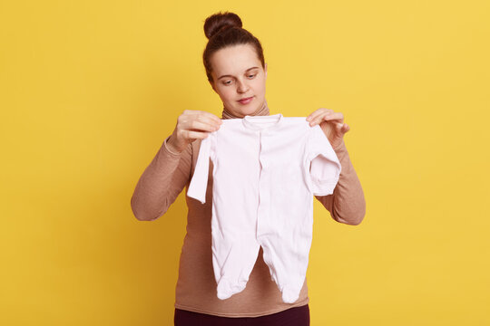 Portrait of beautiful female wearing casual attire, has hair bun, holding newborn sleeper in hands, looking at baby's clothing, ready to giving birth, isolated over yellow background.