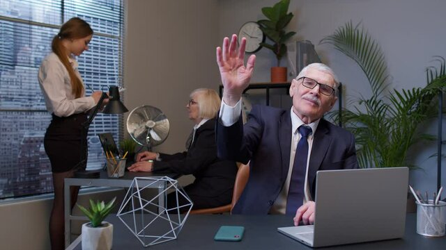 Senior elderly businessman grandfather company director with colleagues team waves palm in hello gesture welcomes someone with hospitable expression expresses positive emotions in office. Coworking