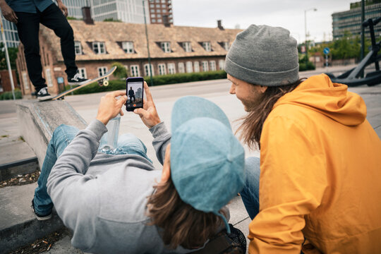 Friends photographing through mobile phone while male skater with skateboard on retaining wall