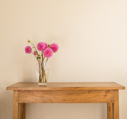 Bright pink dahlias in glass vase on oak side table against beige wall