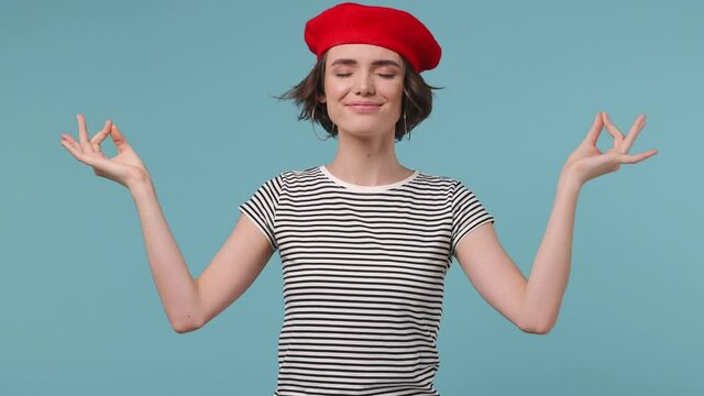 Brunette short hairdo young woman 20s wear red french beret hat striped t-shirt hold spread hands in yoga om gesture relax meditating try to calm down isolated on pastel blue color background studio
