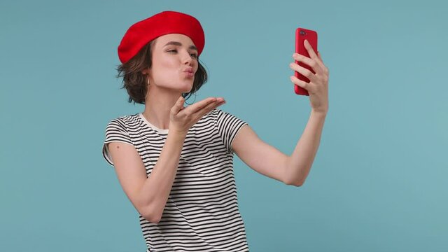 Brunette short hairdo european young woman 20s wear red french beret hat striped t-shirt doing selfie shot on mobile phone post photo on social network isolated on pastel blue color background studio
