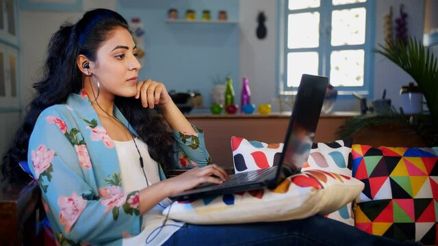 Smart businesswoman working online from home - remote working system. Medium shot of a focused lady sitting on a sofa wearing an earphone while working on a laptop