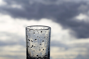 Drinking glass covered with water drops on background of dark sky with storm clouds. Concept of rainy weather