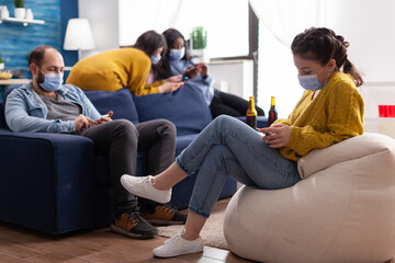 Group of mixed race friends browsing on smartphone keeping social distancing wearing face mask to prevent spread of coronavirus during global pandemic in living room with beer bottle. Conceptual image