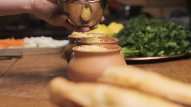 In restaurant kitchen, chef has clay pots on wooden table. cook puts the grated cheese into pots.