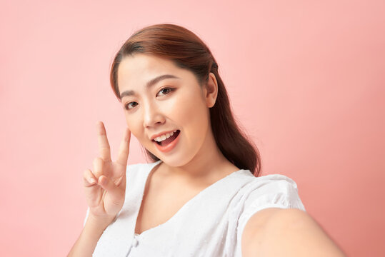 Happy expressive young woman posing while taking pictures of herself through cellphone, over pink background