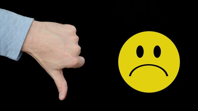 Man showing dislike gesture and unhappy sign. Close-up view of male hand showing thumb down and yellow sad emoticon on black background
