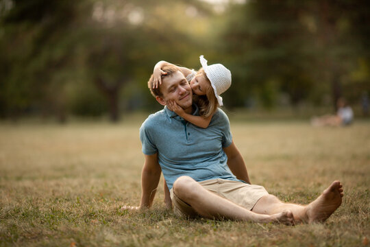 Dad and daughter in summer park sit on the grass. Girl hugs and kisses the dad, father smiles and happy. Family leisure concept, nature walk, family care and values, fatherhood, fathers day