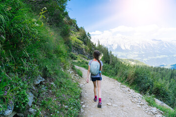 Fototapeta premium Girl hiking on beautiful summer day in alps mountains Austria, resting on rock and admire amazing view to mountain peaks. Active family vacation leisure with kids. Outdoor fun and healthy activity