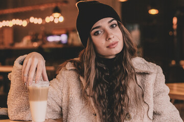 
A beautiful fashionable appearance woman sits at a table in a restaurant holding a latte in her hands and smiles