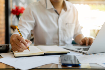 Businessman is writing note and using laptop. Businessman is analyzing financial report on notebook.