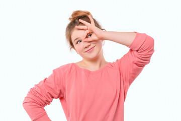 Funny young girl in pink casual sweatshirt, showing OK sign over eye and smiling, having faith all be okay