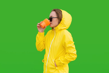 A redhead girl in sunglases and a raincoat drinks coffe from bamboo eco cup and looks away