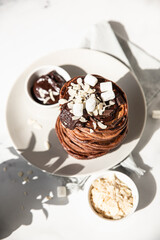 Stack of homemade chocolate pancakes with melted chocolate, ground almond and marshmallow, light background. Direct sunlight and hard shadows.